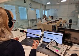 Person med hörlurar sitter i tolkbås med mikrofon och datorskrämar. Genom glasruta syns fler tolkbås