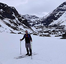 Image of a smiling man on  cross-county skis with a ski pole in each hand making contact with the earth. The man has closely-cropped dark hair and dark, short facial hair. He is wearing blue-toned reflective sunglasses, a long-sleeve black shirt, light gray pants, ski boots,  and a backpack with blue straps. The man is seen standing in his skis on snow with lightly snow-covered, rocky mountains behind him and a light gray, cloudy sky in the background.