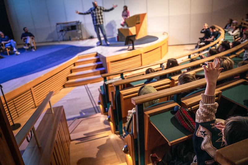 En elev räcker upp en hand. I bakgrunden ser man en scen där en forskare står och pratar.