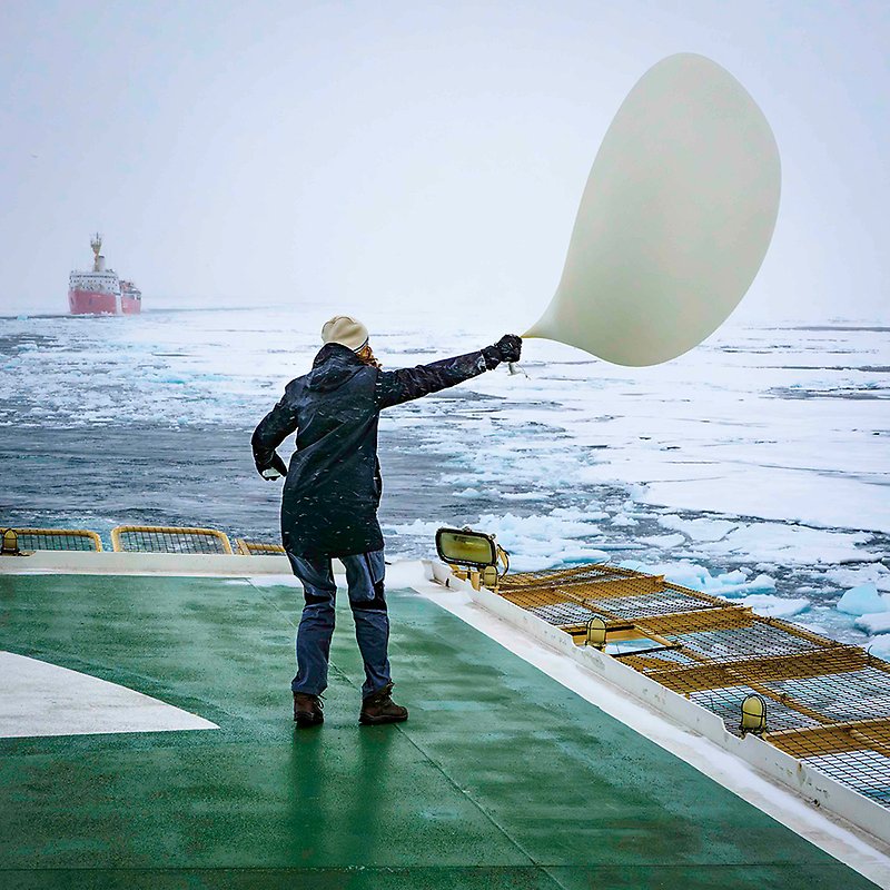 Person standing on deck holding a weather balloon wobbling in the wind. Water, ice and an icebreaker in the far distance, in the background.