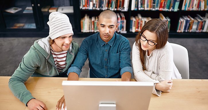 Tre studenter sitter i ett bibliotek och läser på en bildskärm.