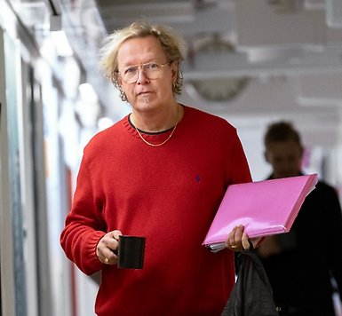 Gunnar Andersson står i en korridor med en kaffekopp i ena handen och en bunt papper i andra.