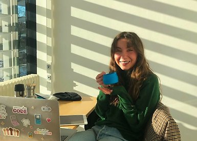 Eleonora Svanberg som håller en kopp kaffe i handen och sitter framför sin dator.