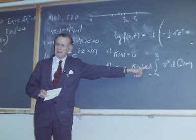 Man i kostym föreläser vid svart tavla med matematiska formler.
