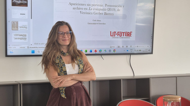 Una mujer con gafas de pie con los brazos cruzados frente a una gran pantalla que muestra una presentación de PowerPoint en español sobre la obra La compañía (2019) de Verónica Gerber Bicecci. La presentación es de Carla Alanís de la Universidad de Estocolmo dentro del proyecto LIT-FUTURE. La sala tiene sillas rojas y una mesa con una jarra de agua.