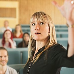 Lärare som föreläser i storsal med studenter i bakgrunden.
