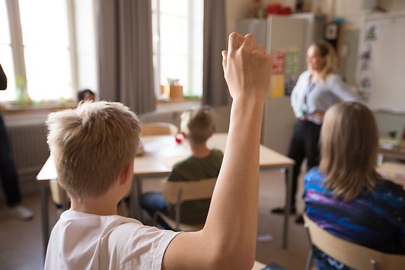 Klassrum. I förgrunden räcker en elev upp handen.