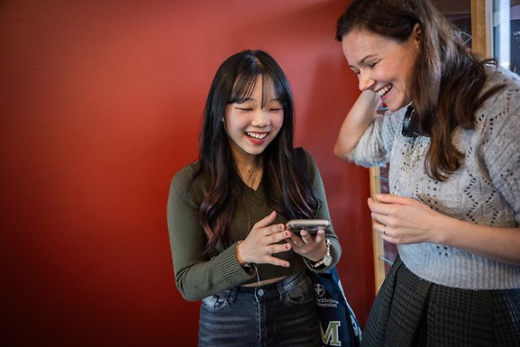 Två studenter med olika kulturella bakgrunder tittar på en mobiltelefon och skrattar.