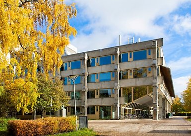 Träd och grönska till vänster. I mitten av bilden är en gångväg som leder till en betongbyggnad. Till höger finns ett träd. 