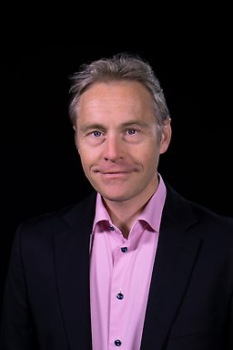 Porträttfoto av Per Carlbring i rosa skjorta och mörk kavaj mot en svart bakgrund.