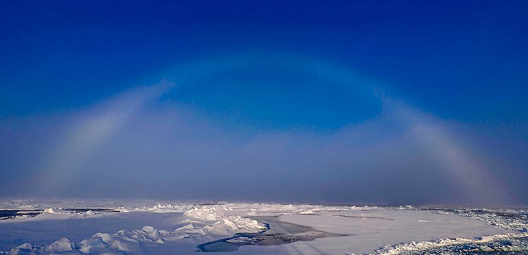 A white fogbow against a clear blue sky, snow and ice.