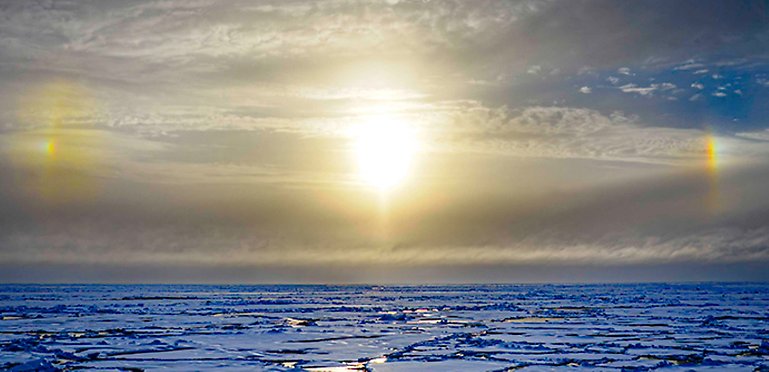 Cloudy sky, blue ice, bright sun, "sundogs" to the left and right, rainbow coloured.