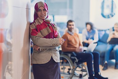 Woman in hijab standilng leaning against a wall. Man sitting in wheelchair on teh background, and people on a couch.