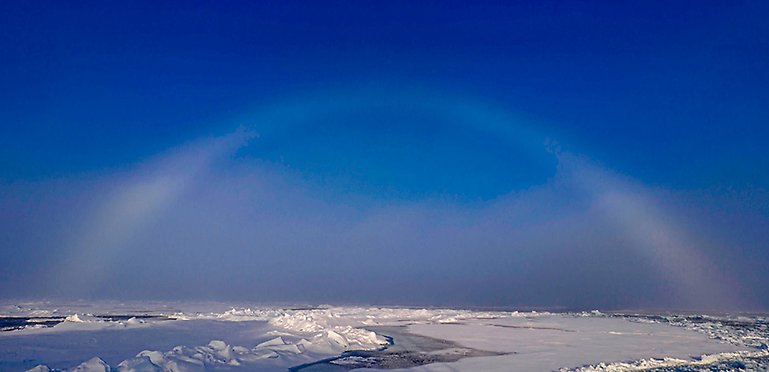 Blå himmel, dimbåge över is och snö.