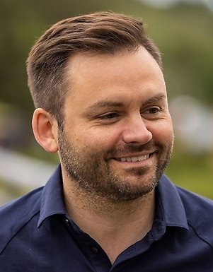 Närbildsporträtt av en person med kort brunt hår och skägg, klädd i en mörkblå skjorta. Bakgrunden är utomhus och mjukt oskarp i gröna toner.