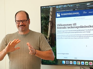 En person pratar teckenspråk. Välkommen till svenskt teckenspråkslexikon står det på en skärm bakom.