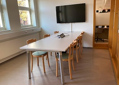 Grupprum med bord och sex stolar samt bildskärm på väggen. 