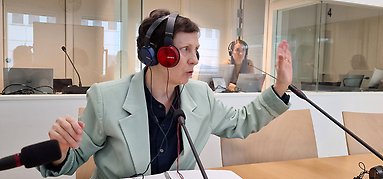 Elisabeth sitter med två par hörlurar på sig vid ett bord med mikrofon och pratar och gestikulerar. I bakgrunden tolkbås.