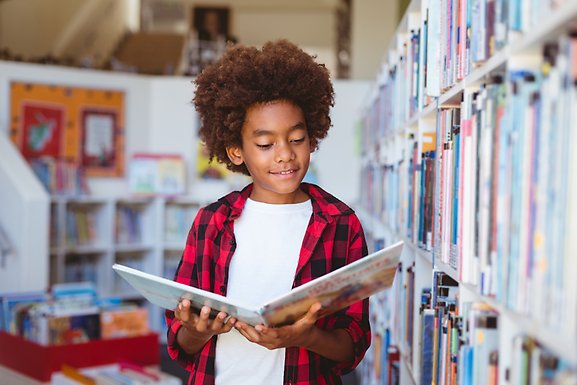 Ett barn står och håller i och läser en bok i ett bibliotek.