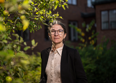 Rebecca Brinch står utomhus bredvid ett äppelträd framför ett brunt tegelhus