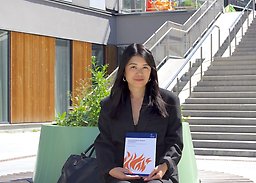 Joanna holding her thesis, sitting on a bench outside on campus Albano.