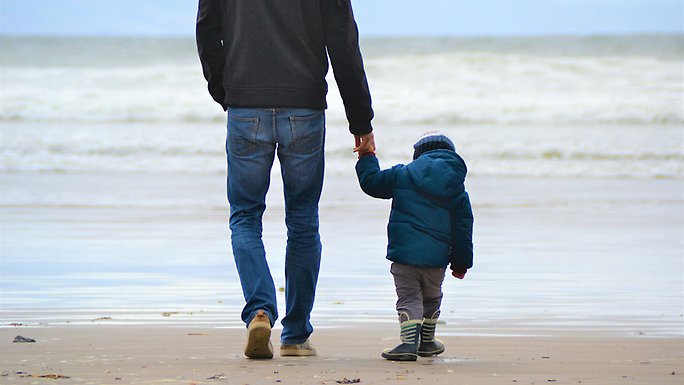 A parent holding a toddler's hand as they walk on a beach.