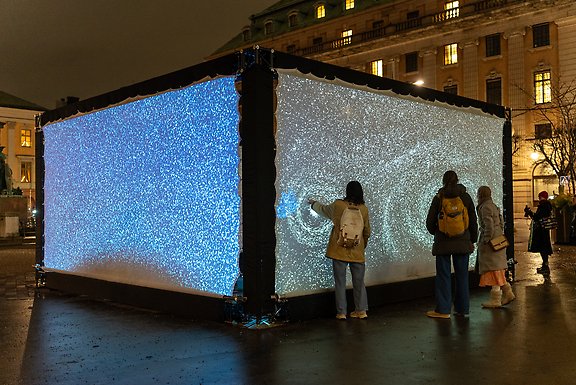 Besökare interagerar med en ljusinstallation som visar virvlande ljusmönster på stora skärmar utomhus.