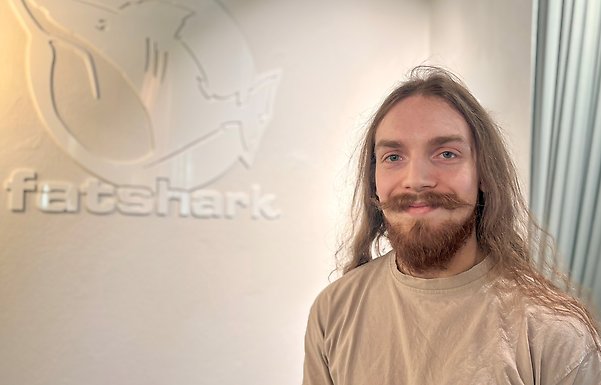 Joel Zetterberg ler och tittar in i kameran. Han står framför en vit vägg där ordet Fatshark syns i bakgrunden.