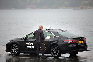 A taxi driver standing next to his car, lake in the bakground.