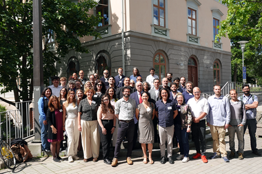 Group photo of numerous participants at the NOLAN 2026 conference organized by NILAS (Nordic Institute of Latin American Studies) at Stockholm University. The participants are gathered in several rows on outdoor steps in front of a light pink and grey building with large windows and arched doorways. The photo was taken on a sunny summer day with green trees in the background.