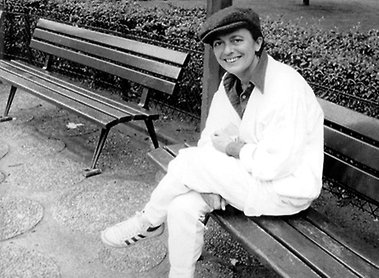 Bilden visar Monique Wittig (1935–2003), den franska författaren, feministiska teoretikern och lesbiska aktivisten, i ett svartvitt porträtt fotograferat av Colette Geoffrey.I bilden sitter Wittig på en parkbänk i en utomhusmiljö, troligen i en park eller trädgård. Hon är klädd i en ljus jacka, ljusa byxor och en mörk keps. På fötterna bär hon Adidas-träningsskor med de karakteristiska tre ränderna. Hon sitter avslappnat med benen i kors och ler mot kameran, vilket ger bilden ett informellt och personligt uttryck. Bakgrunden visar ytterligare bänkar, buskar och ett stenlagt underlag.Fotot fångar en vardaglig, nästan lekfull stämning – en kontrast till hennes intellektuella och revolutionära persona som en av nyfeminismens centrala teoretiker i Frankrike. Wittig är mest känd för sin banbrytande roman Les Guérillères (1969) och sina teorier om heteronormativitet och "könet som klass", som har haft enormt inflytande på feministisk och queer-teori.Bilden är licensierad under CC BY-SA 4.0 och tillgänglig via Wikimedia Commons, publicerad av Succession littéraire de Monique Wittig (Monique Wittigs litterära arv).