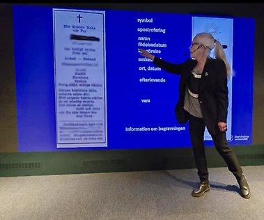 Catrin pekar på en dödsannons som visas på en storbildsskärm.