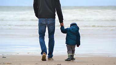 En pappa och hans lilla son går hand ihand på en strand.