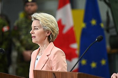 Ursula von der Leyen, ordförande för Europeiska kommissionen, talar vid ett podium under sitt besök i Kanada. Hon bär en ljus persikofärgad kavaj över en vit blus, med ett pärlhalsband. Bakom henne syns den kanadensiska flaggan (röd och vit med ett lönnlöv) och Europeiska unionens flagga (blå med gula stjärnor). En uniformsklädd medlem av de kanadensiska styrkorna står i bakgrunden.
