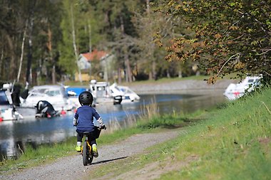 Ett ensamt barn cyklar på en grusväg intill vattnet där segelbåtar är förtöjda.