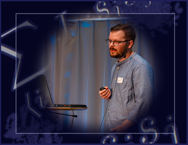 An image of Marcus standing on a stage holding a laser pointer in front of a computer while speaking to a crowd.