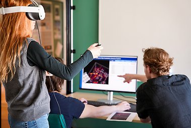Visiting researcher Olivia Mostow tries IDaVIE during a session at the Oskar Klein Centre, with master's student Florian Unger pointing out a structure on the desktop monitor. The screen shows the Manticore reconstruction of a patch of the local universe; the controller is painting directly into that volume inside the headset.