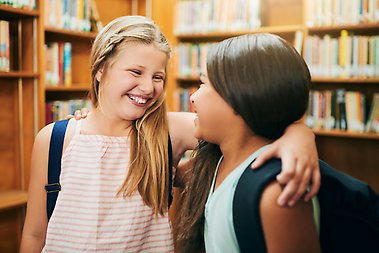 Two young schoolgirls standing with their arms around each other inside a school library during the day