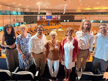 A group of seven people standing in a row inside the European Parliament hemicycle in Brussels, with rows of orange seats and the EU flag visible in the background. The group includes four women and three men dressed in business-casual attire, most wearing name tags, posing for a photo on the upper level overlooking the debating chamber.