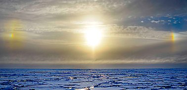 Cloudy sky, blue ice, bright sun, "sundogs" to the left and right, rainbow coloured.