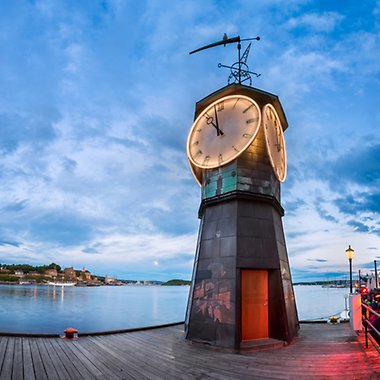 Klocktorn som står på Aker brygge vid vatten och hus i bakgrunden.