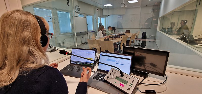 Person with headphones on, viewed from the rear at an angle, sitting in an interpretation boot, with microphone and computer screen.