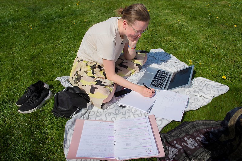 A person sits on a blanket on a green lawn studying outdoors. The person is writing in a notebook with a pen and has a laptop, notebooks, and papers spread out in front of them. The person has light brown hair tied up in a bun, wears glasses, a beige t-shirt, and a patterned skirt in yellow, black, and beige tones. To the left of the person lies a pair of black sneakers and a black bag. It is a sunny day with yellow dandelions in the grass.