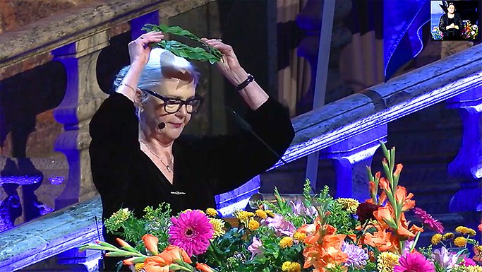 Catrin Norrby i svart klänning sätter på sig lagerkrans. Står vid podium smyckat av blommor.
