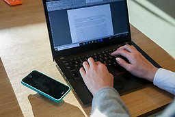 Telefon och laptop på ett bord, och en persons händer som skriver på laptopen.