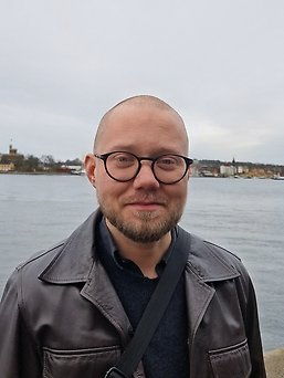 Närbildsporträtt av en person med kortklippt hår, skägg och runda mörka glasögon, klädd i mörk jacka med en axelrem över bröstet. I bakgrunden syns vatten och bebyggelse längs en strand under en mulen himmel.