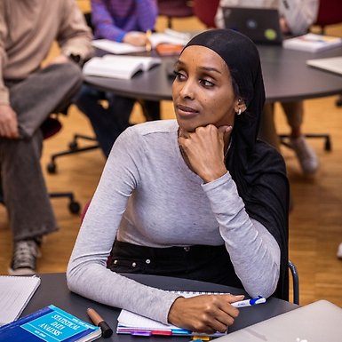 Kvinna sitter vid ett bort och lutar sig mot handen. Statistikböcker och laptop finns på bordet.