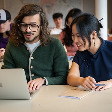 Studenter i ett klassrum. En glad man och en glad kvinna tittar på mannens laptop i förgrunden.