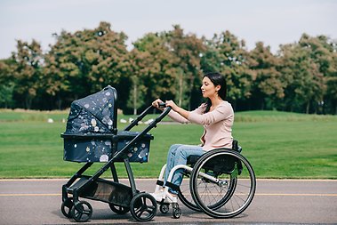 Young disabled woman in wheelchair walking with baby carriage in park.