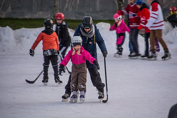 Pappa hjälper dotter styra en hockeyklubba på isen. Andra föräldrar och barn i bakgrunden.